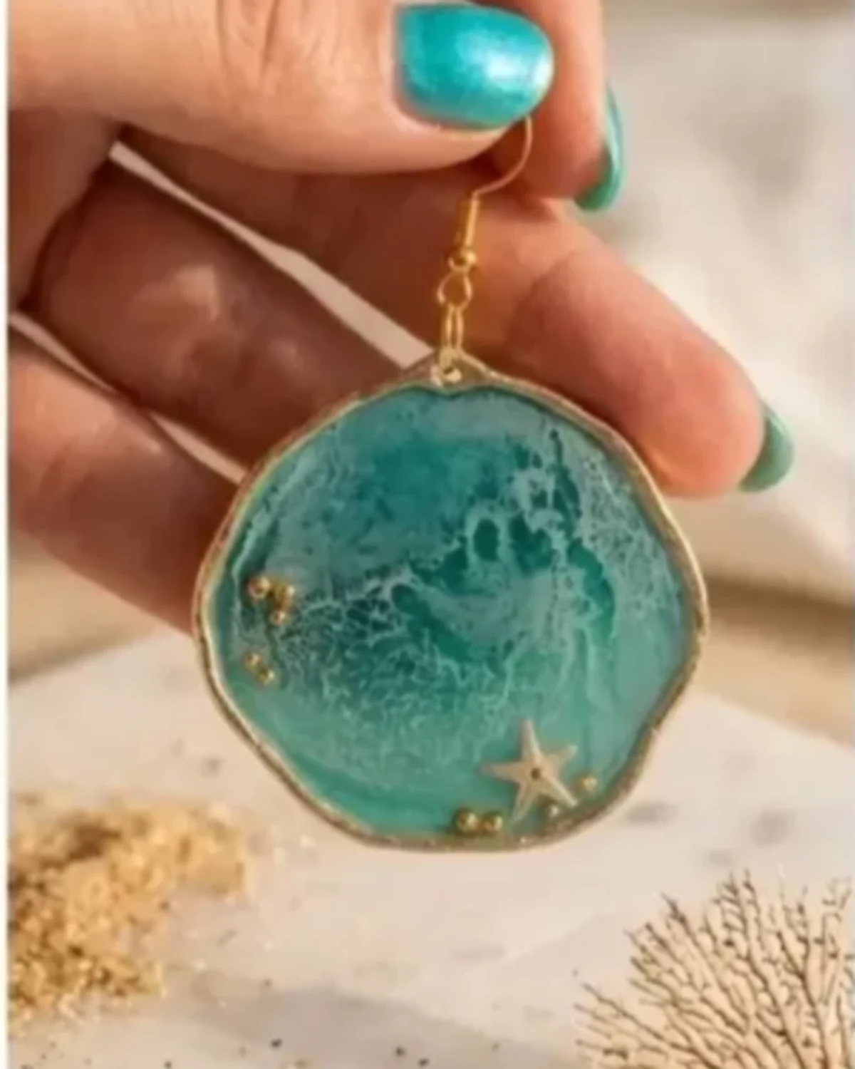 Round teal ocean resin earring with gold edging, a small starfish and gold beads, hanging against sand and coral